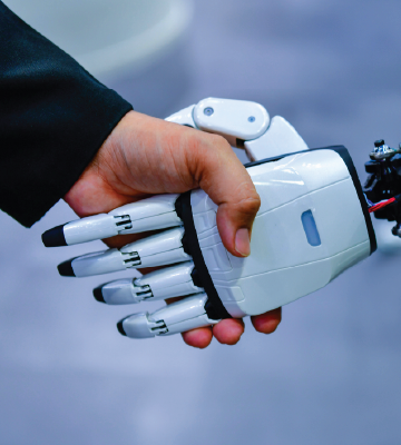 A human hand in a dark suit sleeve shakes hands with a white robotic hand. The robotic hand has segmented fingers and a smooth, plastic-like surface. The background is softly blurred in shades of blue and gray.