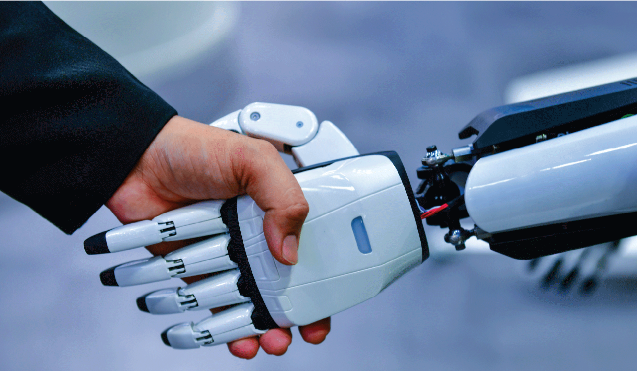 A human hand in a dark suit sleeve shakes hands with a white robotic hand. The robotic hand has segmented fingers and a smooth, plastic-like surface. The background is softly blurred in shades of blue and gray.