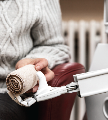A person in a ribbed sweater sits in a chair, holding a rolled bandage. A robotic arm extends from the right, reaching toward the bandage. The background shows a radiator and part of a wall.