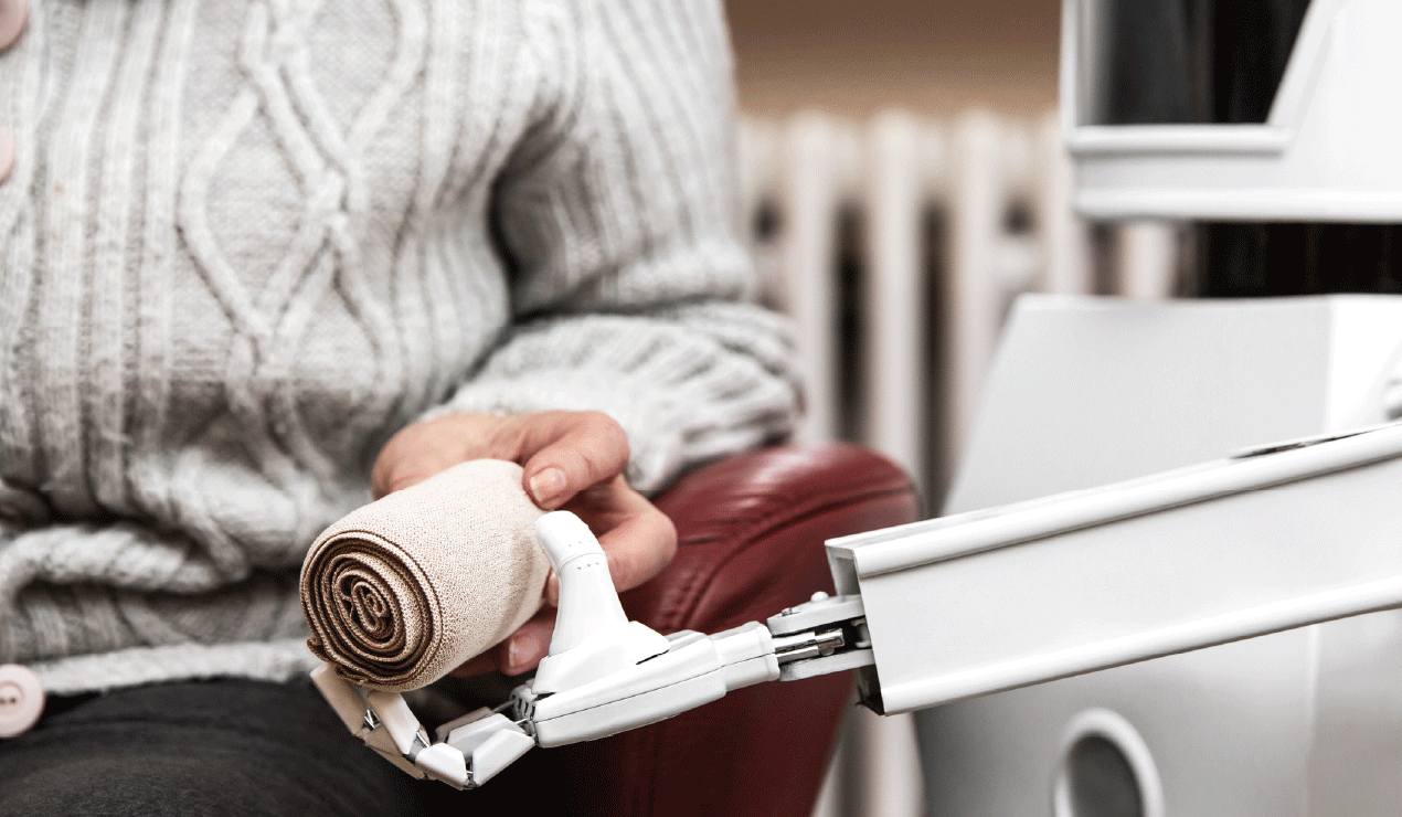 A person in a ribbed sweater sits in a chair, holding a rolled bandage. A robotic arm extends from the right, reaching toward the bandage. The background shows a radiator and part of a wall.