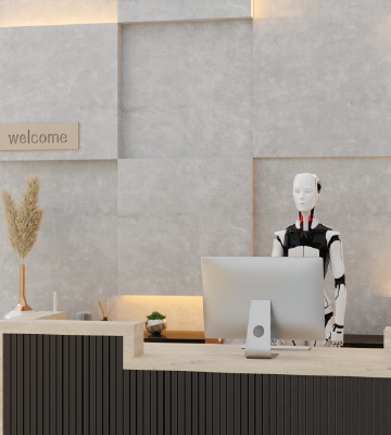 A humanoid robot stands behind a modern reception desk, facing a large computer monitor. The desk has a light wood top and dark vertical paneling. Decorative branches and a vase with dried grass are placed on the counter. The wall behind the desk is light gray with geometric paneling, and a sign with the word 'welcome' is mounted on the left side.