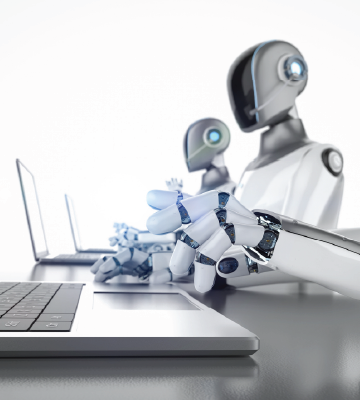 Humanoid robots with smooth, metallic surfaces sit at a row of desks, each using a laptop. The robots have articulated fingers and featureless faces, with a streamlined, futuristic design. The background is plain and bright, emphasizing the robots and their workstations.