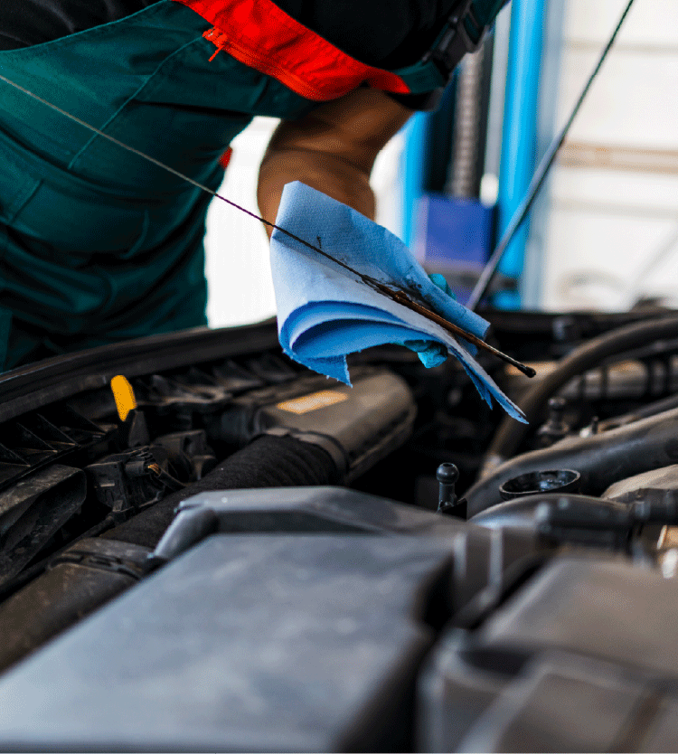 Person in green overalls and red shirt holding a blue cloth and cleaning an engine compartment of a vehicle