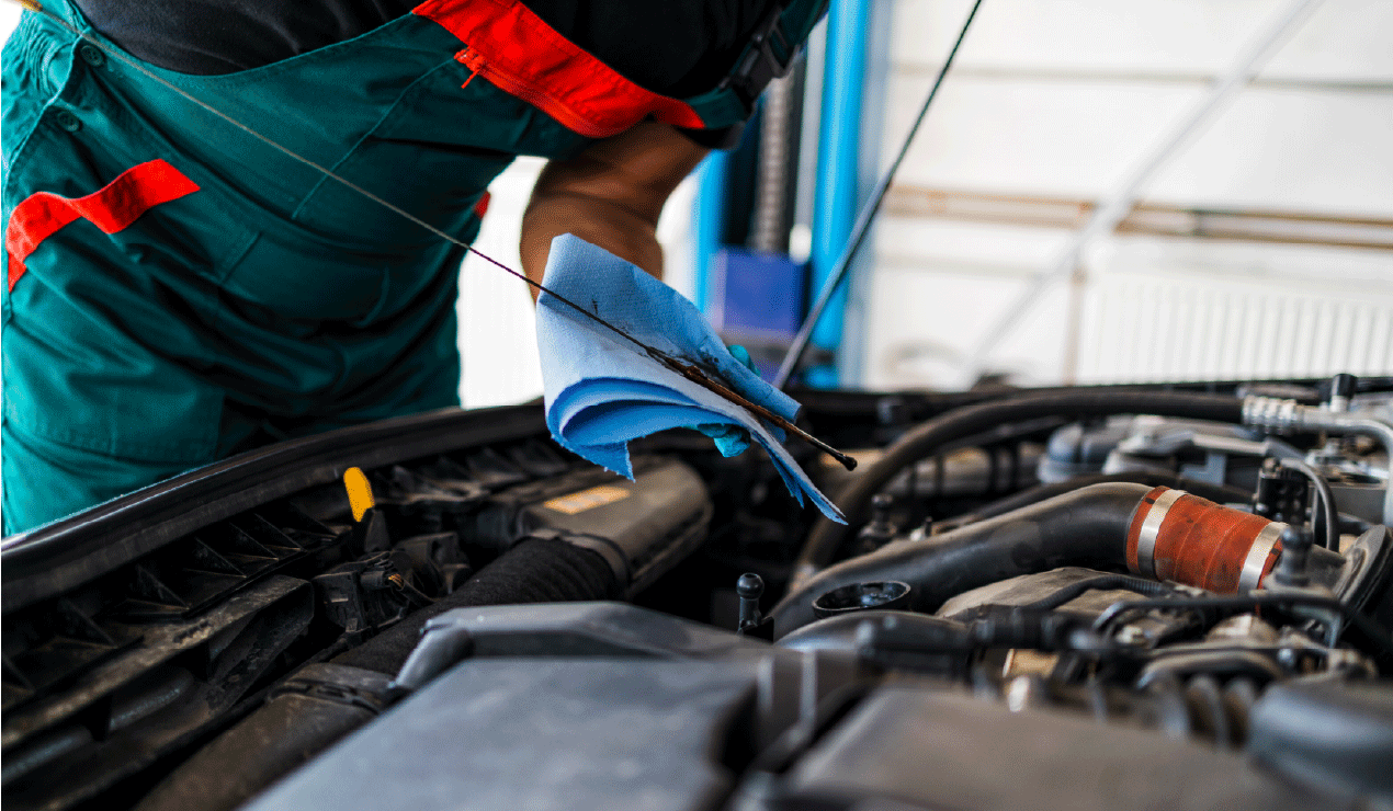 Person in green overalls and red shirt holding a blue cloth and cleaning an engine compartment of a vehicle