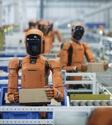 Orange humanoid robots stand in rows along a conveyor system inside a large industrial facility. Each robot holds a rectangular box and faces forward. The background shows shelves, bins, and additional robots, suggesting an automated assembly or packaging environment. The lighting is bright and even, highlighting the organized, repetitive layout of the workspace.