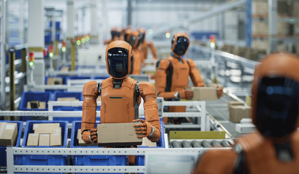 Orange humanoid robots stand in rows along a conveyor system inside a large industrial facility. Each robot holds a rectangular box and faces forward. The background shows shelves, bins, and additional robots, suggesting an automated assembly or packaging environment. The lighting is bright and even, highlighting the organized, repetitive layout of the workspace.